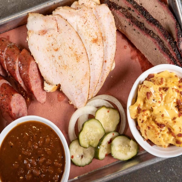 A BBQ platter with sliced turkey, brisket, sausage, baked beans, mac and cheese, pickles, and onions.